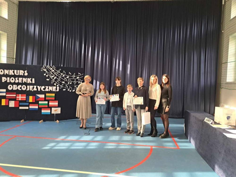 Zdjęcie przedstawia uczestników konkursu piosenki obcojęzycznej z nauczycielami w tle tytuł konkursu i flagi różnych państw